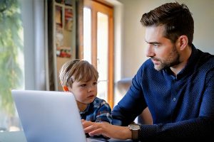 Ein Vater arbeitet am Laptop, während sein kleines Kind neben ihm am Fenster steht
